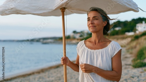 Mature woman holding a large beach umbrella while standing on sandy shore near the ocean, with coastal houses and greenery visible in the background