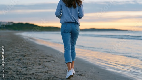 Female walking along sandy beach at sunset, wearing casual blue sweater and jeans, with ocean waves gently lapping at the shore in the background