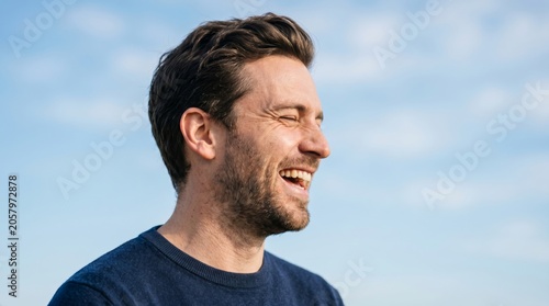 A joyful Caucasian man with short brown hair smiles brightly against a clear sky backdrop.