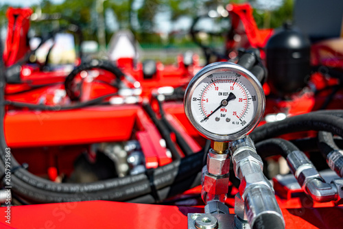 Detailed close-up view of a round pressure gauge mounted on a metal hydraulic manifold, surrounded by black hydraulic hoses and bright red painted machinery