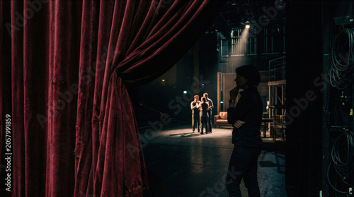 Theater director watching actors rehearse on stage behind a red curtain in a dark playhouse.