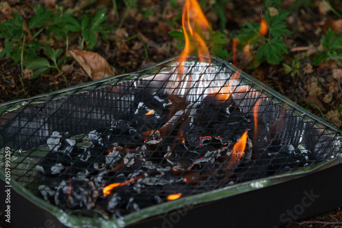 Burning charcoal glows brightly in a disposable grill over leaves and dirt.