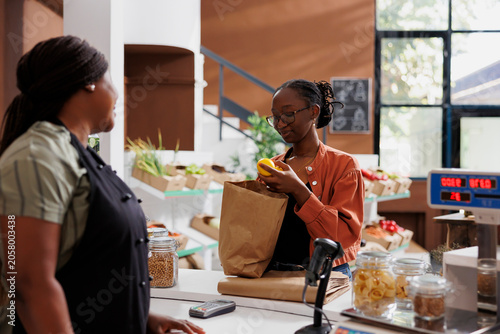 African American woman vendor at a small grocery store with a female customer, promoting healthy and sustainable produce. Young client with brown paper bag, unloading fresh lemons at counter.