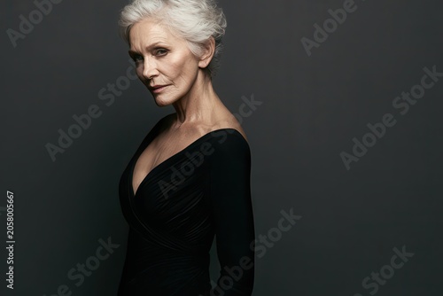 Sophisticated mature woman with silver hair posing confidently in a black dress, photographed in a studio portrait celebrating elegance, beauty and graceful aging.