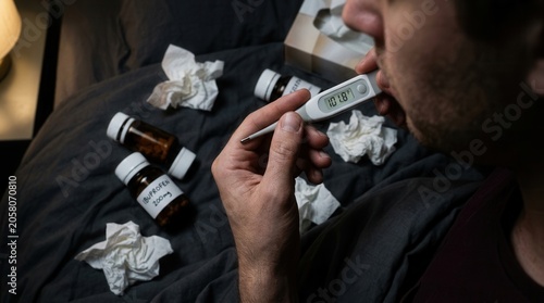 Man Taking Temperature While Sick In Bed With Medicine And Tissues