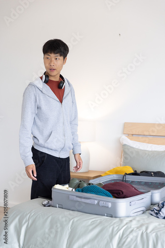 Asian man standing beside bed in bedroom, packing silver suitcase with headphones, copy space
