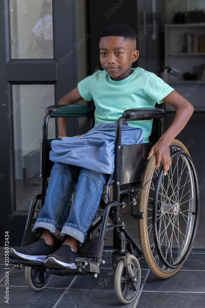 Obraz premium African American schoolboy sitting in manual wheelchair at entrance gripping wheel, backpack on lap