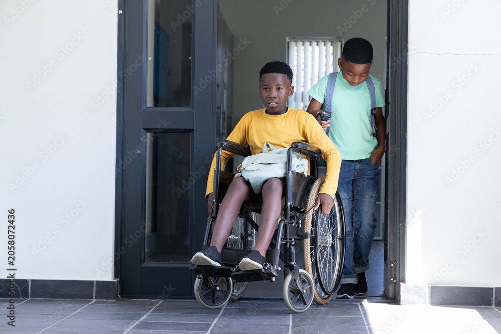 Obraz premium African American school-age boys pushing manual wheelchair through dark door at entrance, backpacks