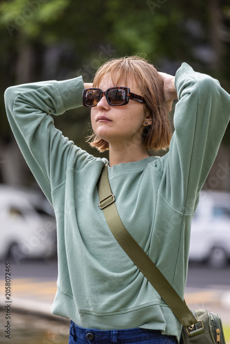 Woman standing with hands behind head on leafy street, wearing green crossbody bag and sunglasses