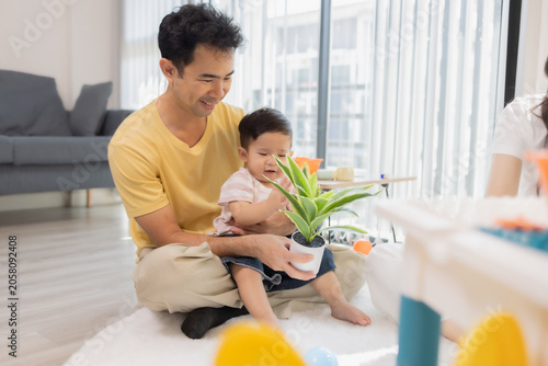 
Warm sunlight streams through blinds. Family of three smiles softly in cozy room. Mother holds infant close, father looks lovingly on. Green plant adds freshness to minimalist shelves
