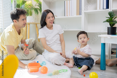 
Warm sunlight streams through blinds. Family of three smiles softly in cozy room. Mother holds infant close, father looks lovingly on. Green plant adds freshness to minimalist shelves
