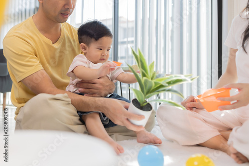 
Warm sunlight streams through blinds. Family of three smiles softly in cozy room. Mother holds infant close, father looks lovingly on. Green plant adds freshness to minimalist shelves
