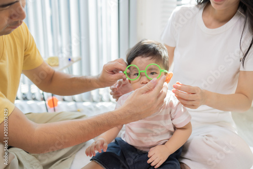 
Warm sunlight streams through blinds. Family of three smiles softly in cozy room. Mother holds infant close, father looks lovingly on. Green plant adds freshness to minimalist shelves
