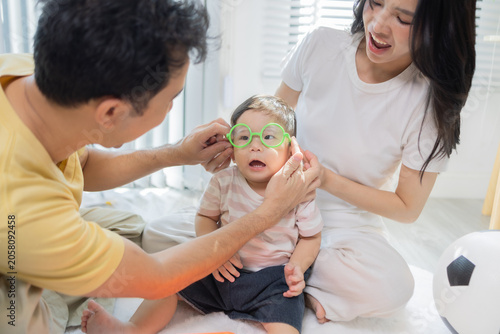 
Warm sunlight streams through blinds. Family of three smiles softly in cozy room. Mother holds infant close, father looks lovingly on. Green plant adds freshness to minimalist shelves
