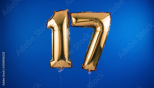 Golden balloons display the number seventeen against a blue backdrop indoors.