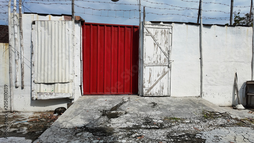 Weathered entrance featuring a striking red door among rustic white walls and cracked concrete groun
