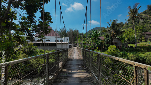 A wooden suspension bridge surrounded by lush greenery leads to a tranquil village setting under a bright blue sky.