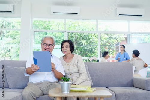 Fotografie Elderly couple receives tablet assistance from attentive nurse during bright, calm moment within comfortable nursing home room featuring expansive window views of vibrant greenery