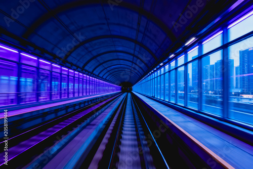 A perspective view of a modern, illuminated subway tunnel with tracks extending into the distance, flanked by glass walls showing an urban cityscape.