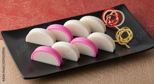 Slices of kamaboko fish cake served on a black rectangular plate with decorative mizuhiki ornaments