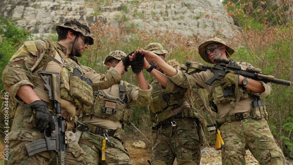 Fototapeta premium A diverse team of soldiers in full combat gear stacks hands together, demonstrating unity and strategizing for a military operation in a rugged environment.