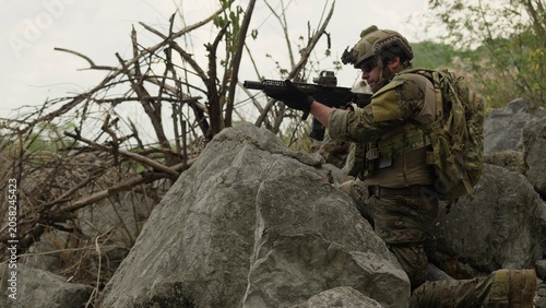 Принти на полотні A special forces soldier conducts a reconnaissance mission, aiming his rifle with focused determination while taking cover behind rocks in rugged, hostile terrain