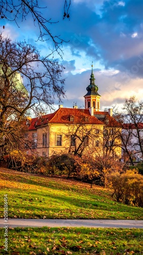 A serene landscape featuring a historic building with a red-tiled roof and a tower, surrounded by trees on a grassy hill under a cloudy sky