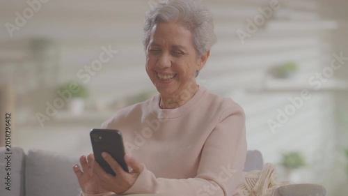elderly Asian woman relaxing on sofa with phone in living room. Connect internet at home Wi-Fi, play social media. transfer money online or financial payment, smile or laugh with happy and positive.