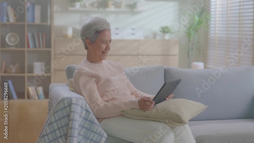 elderly Asian woman relaxing on sofa with tablet in living room. Connect internet at home Wi-Fi, play social media. transfer money online or financial payment, smile or laugh with happy and positive.