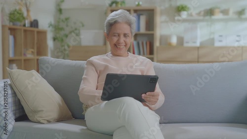 elderly Asian woman relaxing on sofa with tablet in living room. Connect internet at home Wi-Fi, play social media. transfer money online or financial payment, smile or laugh with happy and positive.