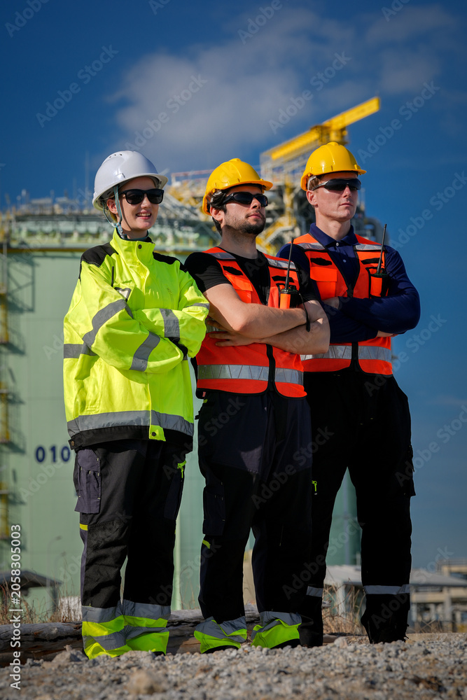 Fototapeta premium Construction worker hard hat safety vest high visibility industrial site teamwork engineer protective eyewear during inspection at refinery with crane, confident stance under blue sky