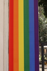 Close-up of a vertical rainbow pride flag hanging between walls, showing vivid stripes of red, orange, yellow, green, blue and violet in an urban outdoor setting.