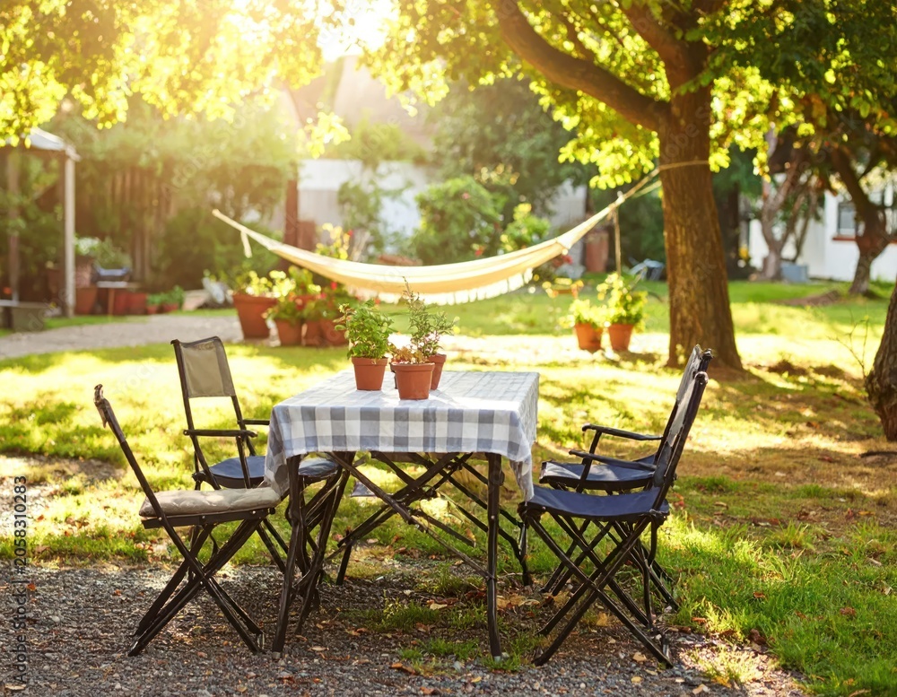 Fototapeta premium Cozy outdoor scene with a table, chairs, hammock, and greenery bathed in sunlight