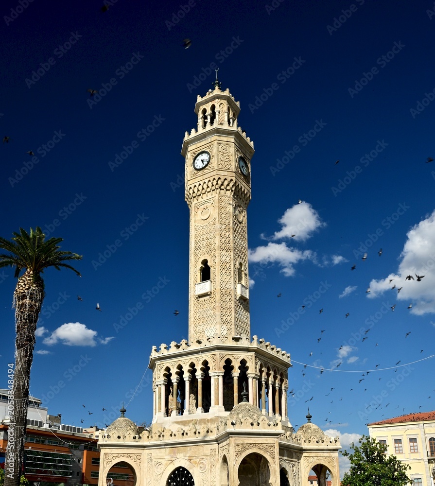 Fototapeta premium Historic Izmir Clock Tower with Pigeons in Flight Against Deep Blue Sky, Konak Square, Turkey
