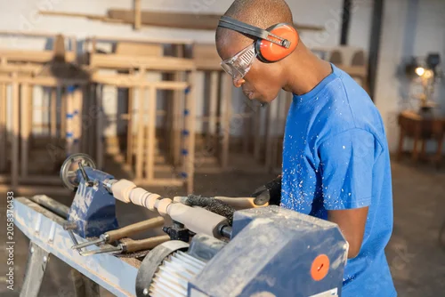 Fototapeta Woodworker shaping cylindrical wood blank on lathe in carpentry workshop, wood shavings scattering