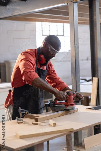 Fototapeta Red power sander smoothing long wooden plank on worktable inside workshop, with wood offcuts