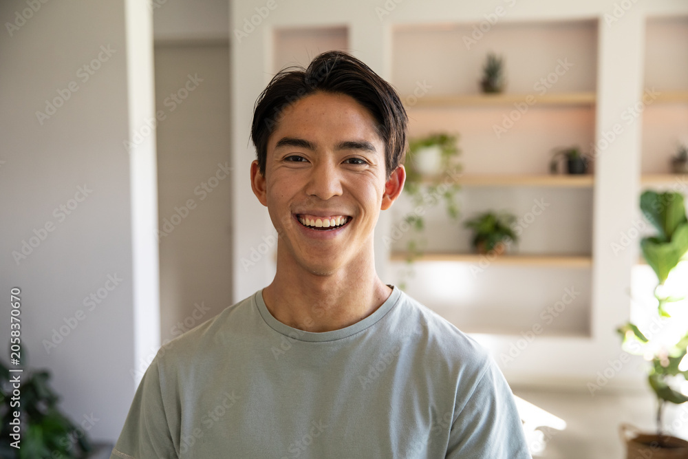 Fototapeta premium Asian man smiling and standing in living room near recessed wooden shelves and plants
