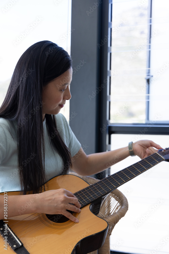 Fototapeta premium Mature African woman tuning wood cutaway guitar, sitting on chair by window, wearing watch
