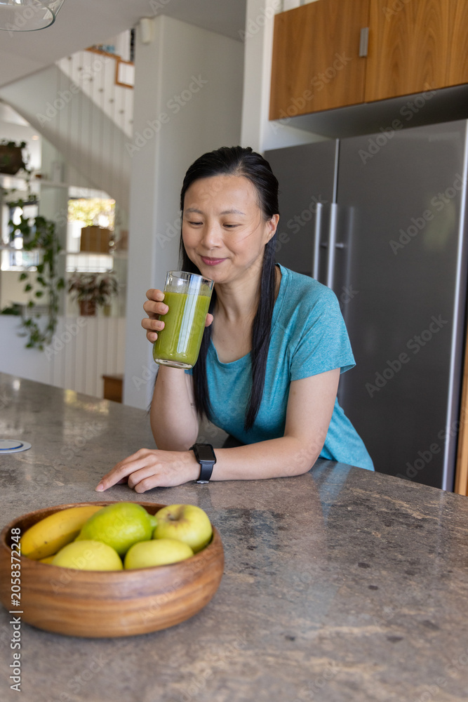 Obraz premium Mature Asian woman standing in kitchen holding tall glass with green smoothie, wearing smartwatch