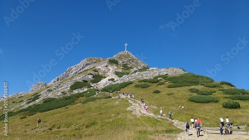 A group of tourists hiking up a rocky stone path towards the summit of Mount Giewont with its iconic steel cross, located in the Tatra National Park near Zakopane, Poland.