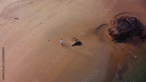 Solitary person is navigating rocky coast. Secluded shoreline with lone wanderer among stones. Male traveler runs along rough coast with shallow green water and textured sand surfaces filmed by drone