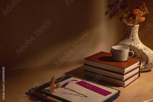 A reading table with books, notebooks, pens, coffee, a vase, and dried flowers creates a wonderful decorative piece with a combination of light, shadow, and color.