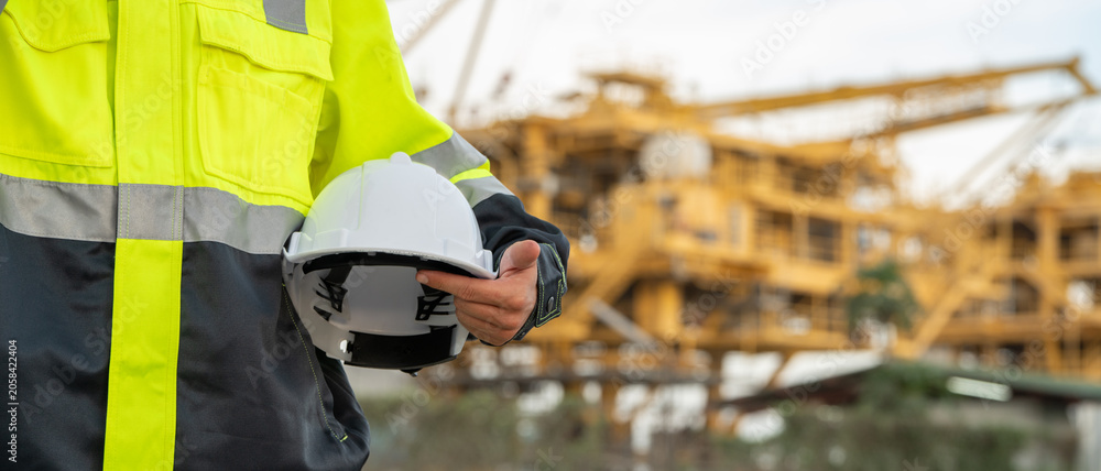 Fototapeta premium Industrial engineer in a high-visibility safety jacket and earmuffs, holding a hard hat and walkie-talkie radio at an offshore energy platform facility. Concept of workplace safety