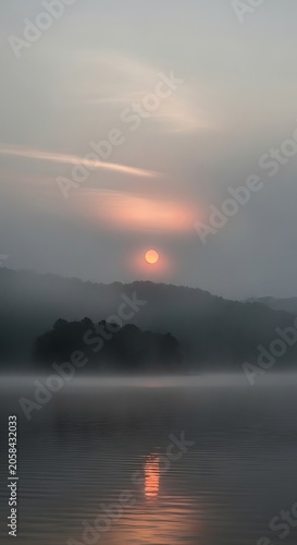 Misty Orange Sun Setting Over Forested Hills and Calm Lake