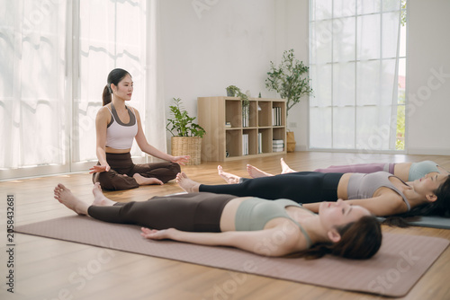 Group of young peaceful Asian women in activewear sitting on yoga mats practicing mindfulness meditation with eyes closed in home studio. Concept of healthy lifestyle and longevity.
