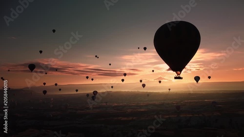 Silhouetted hot air balloons against sunrise sky filmed by drone in Cappadocia. Low angle capture emphasizes single balloon in foreground with soft sun flare and distant convoy. Travelling concept