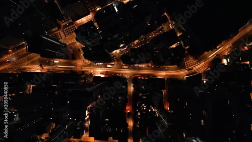 Aerial night artery of illuminated crossroad with golden traffic streams weaving between highrise blocks with neon reflections on asphalt. Dense urban fabric and metropolitan rhythm filmed from above