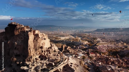 Aerial view of hot air balloons soaring above ancient fortress and rocky valley in Cappadocia at dawn. Colorful envelopes are casting shadows on carved stone formations. Pilot baskets across Anatolia