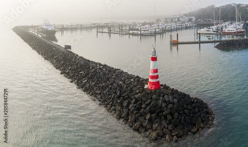 Картината върху платно A red and white lighthouse stands on a rocky breakwater at Orakei Marina, Auckland, New Zealand, guiding boats through the morning fog