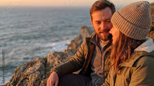 Coastal couple in knit hat at sunset on rocky shore by calm sea, scenic ocean cliff with warm light and sea view